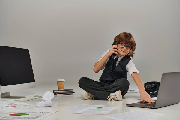 kid in glasses and formal wear talking on smartphone and sitting surrounded by office equipment