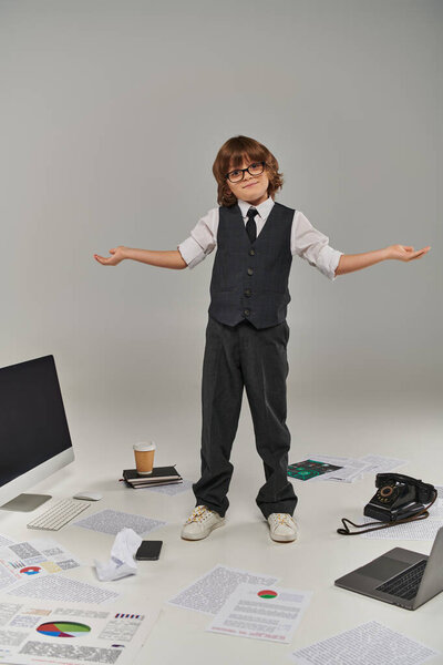 confused kid in glasses and formal wear surrounded by office equipment and devices standing on grey