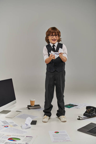 excited boy in glasses and formal wear surrounded by office equipment and devices holding papers