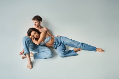 Saten sutyenli esmer genç manken gri arka planda mavi kot pantolonlu seksi üstsüz adama yaslanıyor.
