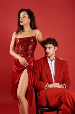 Seksi elbiseli güzel bir kadın kırmızı arka planda resmi giyinmiş bir adamın yanında duruyor, sofistike.