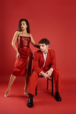 Kırmızı arka planda resmi giyinmiş erkeğin yanında duran seksi elbiseli zarif genç kadın, sofistike.