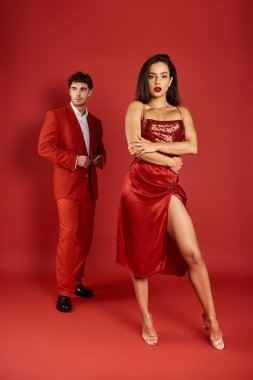 Kırmızı arka planda bulanık bir adamın yanında parlak seksi bir elbise giymiş güzel ve genç bir kadına odaklan.