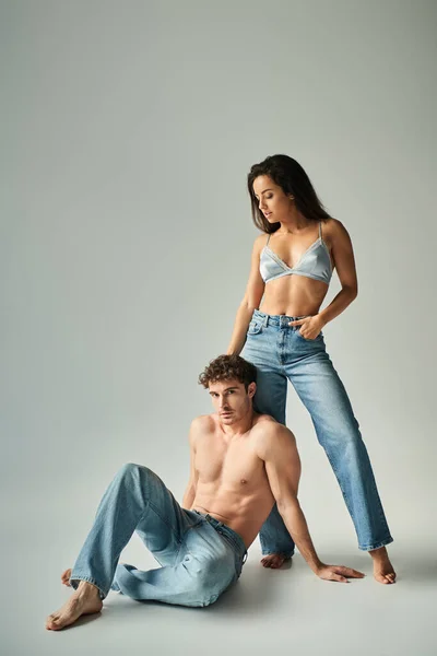 stock image pretty young woman in satin bra and jeans posing near shirtless man on grey background, closeness