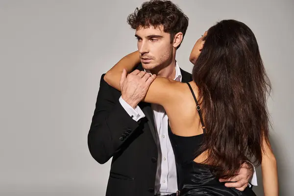 brunette young woman in black slip dress embracing handsome man in evening wear on grey background