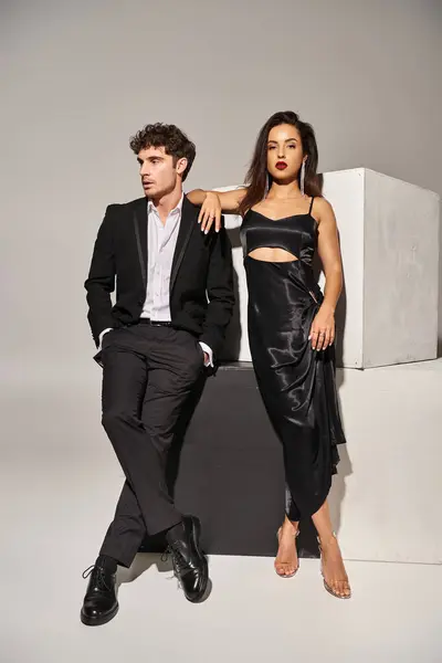 stock image man in suit posing with hands in pockets and looking at gorgeous brunette woman on grey backdrop