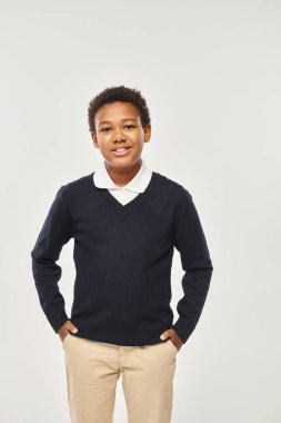 Mutlu Afro-Amerikan okul çocuğu şık bir üniforma içinde elleri gri ceplerinde duruyor.