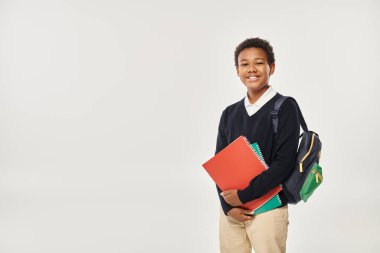 happy african american schoolboy in uniform holding notebooks and standing on grey background clipart
