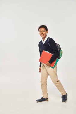 Mutlu Afro-Amerikan okul çocuğu elinde defterlerle gri arka planda duruyor.