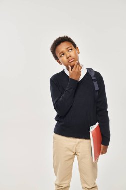 pensive african american schoolboy in uniform holding notebooks and standing on grey background clipart