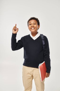 happy african american schoolboy in uniform holding notebooks and showing idea on grey background clipart