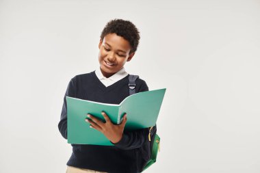 happy african american schoolboy in uniform looking at notebook and standing on grey background clipart