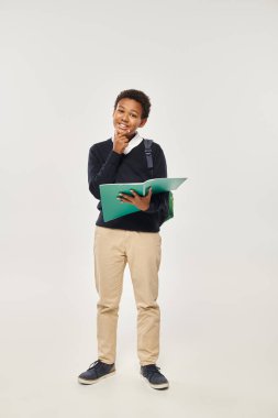 happy african american schoolboy in uniform holding notebook on grey background, hand near chin clipart