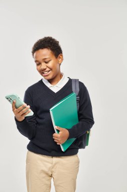Mutlu Afro-Amerikan okul çocuğu gri arka planda akıllı telefon ve defter tutuyor.