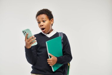 Şok olmuş Afro-Amerikan okul çocuğu gri arka planda akıllı telefon ve defter tutuyor.