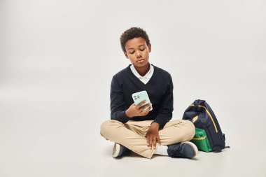 african american schoolboy in uniform using smartphone and sitting near backpack on grey backdrop clipart