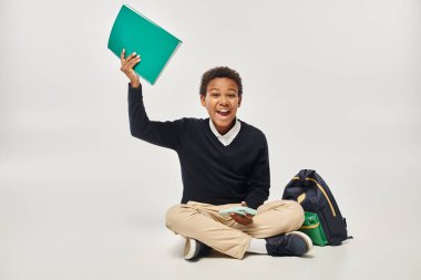 happy african american boy in uniform holding smartphone and notebook while sitting near backpack clipart