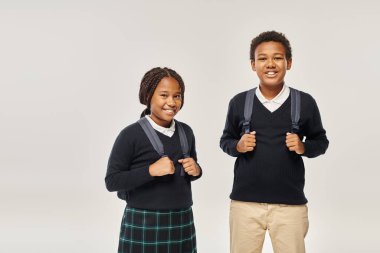 Sırt çantalı mutlu Afro-Amerikan okul çocukları gri arka planda düzgün süveterler giyiyorlar.