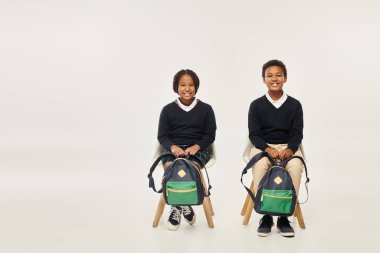cheerful african american schoolkids with backpacks sitting and looking at camera on grey backdrop clipart