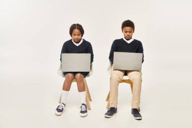 concentrated african american schoolkids in uniform using laptops and sitting on grey backdrop clipart