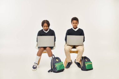 focused african american schoolkids in uniform using laptops and sitting on grey background clipart