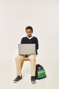 focused african american schoolboy in uniform and glasses using laptop and sitting on grey backdrop clipart