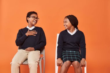 cheerful african american schoolkids in uniform laughing and sitting on chairs on orange backdrop clipart