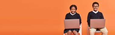 happy african american schoolkids in uniform sitting on chairs and using laptops on orange, banner clipart