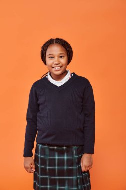 Üniformalı mutlu Afro-Amerikan okul kızı gülümsüyor ve turuncu arka plandaki kameraya bakıyor.