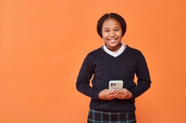 Mutlu Afro-Amerikan okul kızı gülümsüyor ve turuncu arka planda akıllı telefon tutuyor.