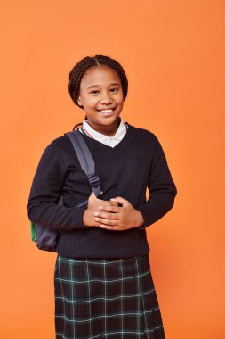 Mutlu Afro-Amerikan okul kızı gülümsüyor ve turuncu arka planda sırt çantası taşıyor.