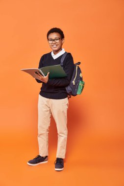 cheerful african american schoolboy in uniform holding backpack and textbook on orange backdrop clipart