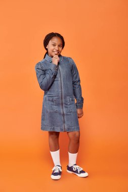 full length of happy african american girl in denim dress posing and smiling on orange backdrop clipart