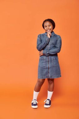 full length of cheerful african american girl in denim dress smiling on orange background clipart