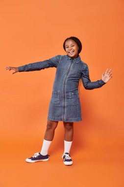 full length of excited african american girl with braids posing in denim dress on orange background clipart