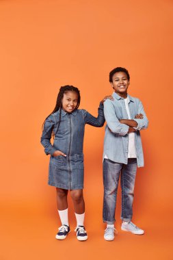 İyimser Afrikalı Amerikalı çocuklar günlük kot giyip turuncu arka planda poz veriyorlar.