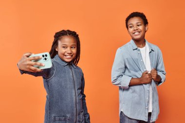 Kotlu elbiseli mutlu Afro-Amerikalı kız turuncu arka planda bir çocukla selfie çekiyor.