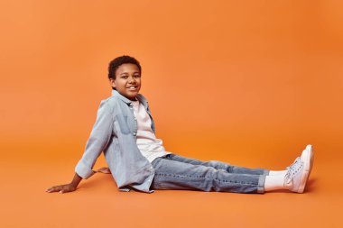 Her gün giyinmiş neşeli Amerikalı çocuk yerde oturuyor ve kameraya gülümsüyor.