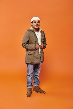 Kışlık ceketli ve bereli neşeli Afro-Amerikan çocuğu kameraya neşeyle gülümsüyor.
