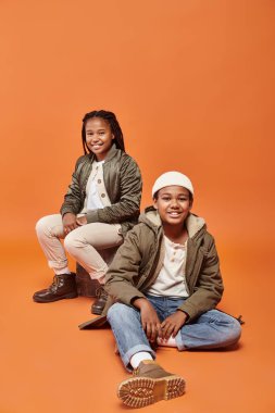 cheerful preteen african american boy and girl in winter attire sitting on floor on ornage backdrop clipart