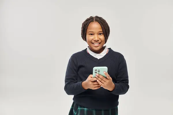 stock image happy preteen african american schoolgirl in uniform holding smartphone on grey background