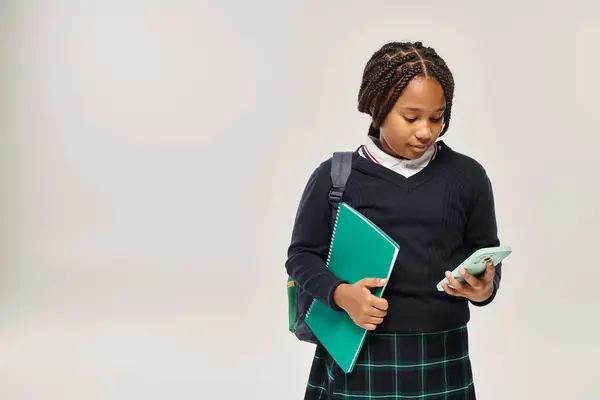 preteen african american schoolgirl in uniform using smartphone and holding notebook on grey