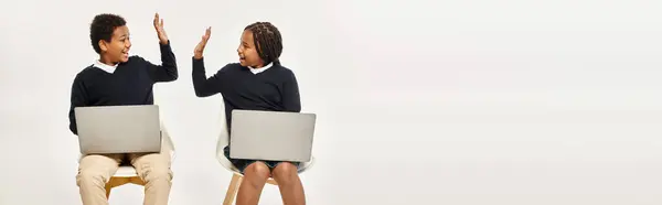 stock image happy african american schoolkids in uniform using laptops and giving high five on grey, banner