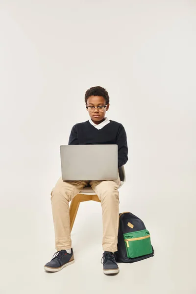 stock image focused african american schoolboy in uniform and glasses using laptop and sitting on grey backdrop