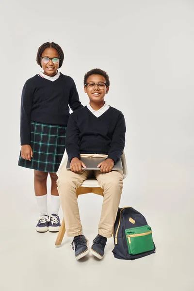 cheerful african american girl in uniform standing near schoolboy with laptop on grey backdrop
