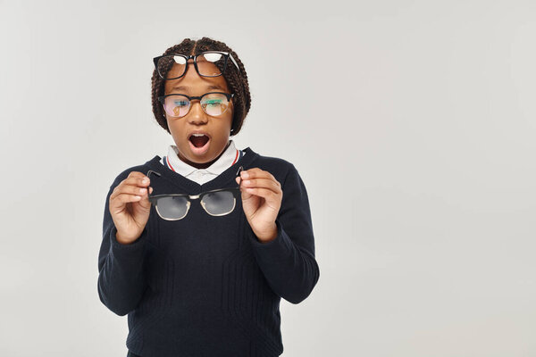 shocked african american girl in sweater and eyewear holding glasses and looking at camera on grey