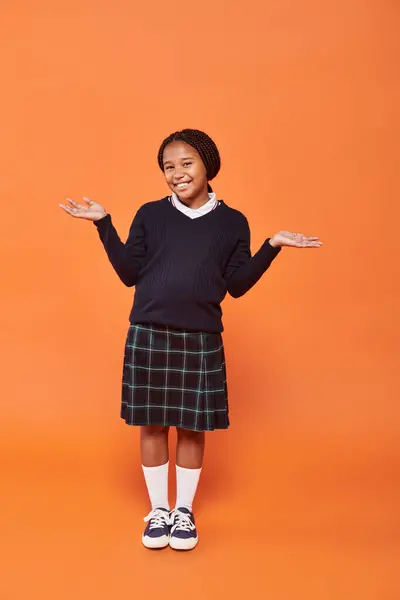 happy african american schoolgirl in uniform gesturing and looking at camera on orange background