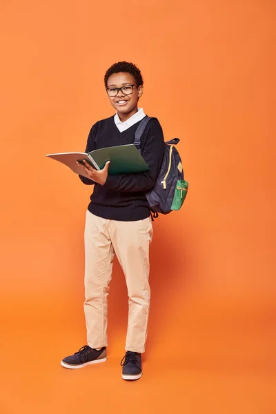 cheerful african american schoolboy in uniform holding backpack and textbook on orange backdrop