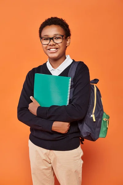 relaxed african american schoolboy in uniform holding backpack and textbook on orange backdrop
