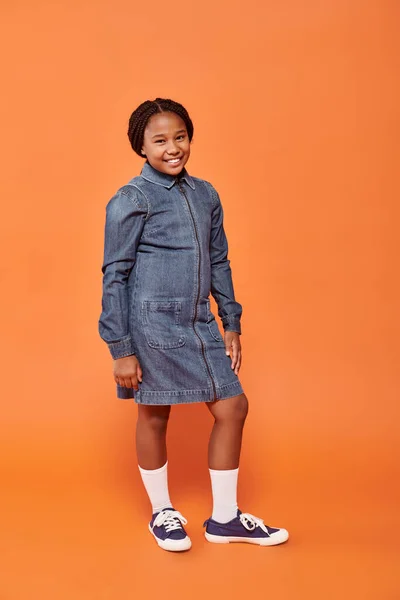 Stock image full length of cheerful african american girl in denim dress posing and smiling on orange backdrop